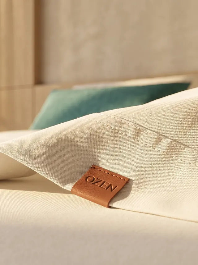 Juego de sábanas OZEN en algodón crema sobre una cama, con la etiqueta de cuero de la marca a la vista